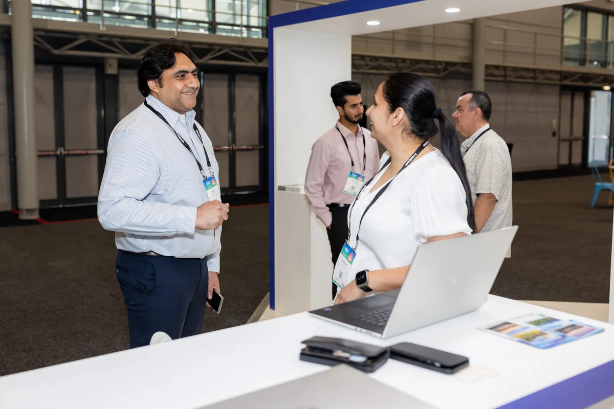 People interact at a booth during an indoor convention, showcasing Sukh Sandhu's work in compliance.
