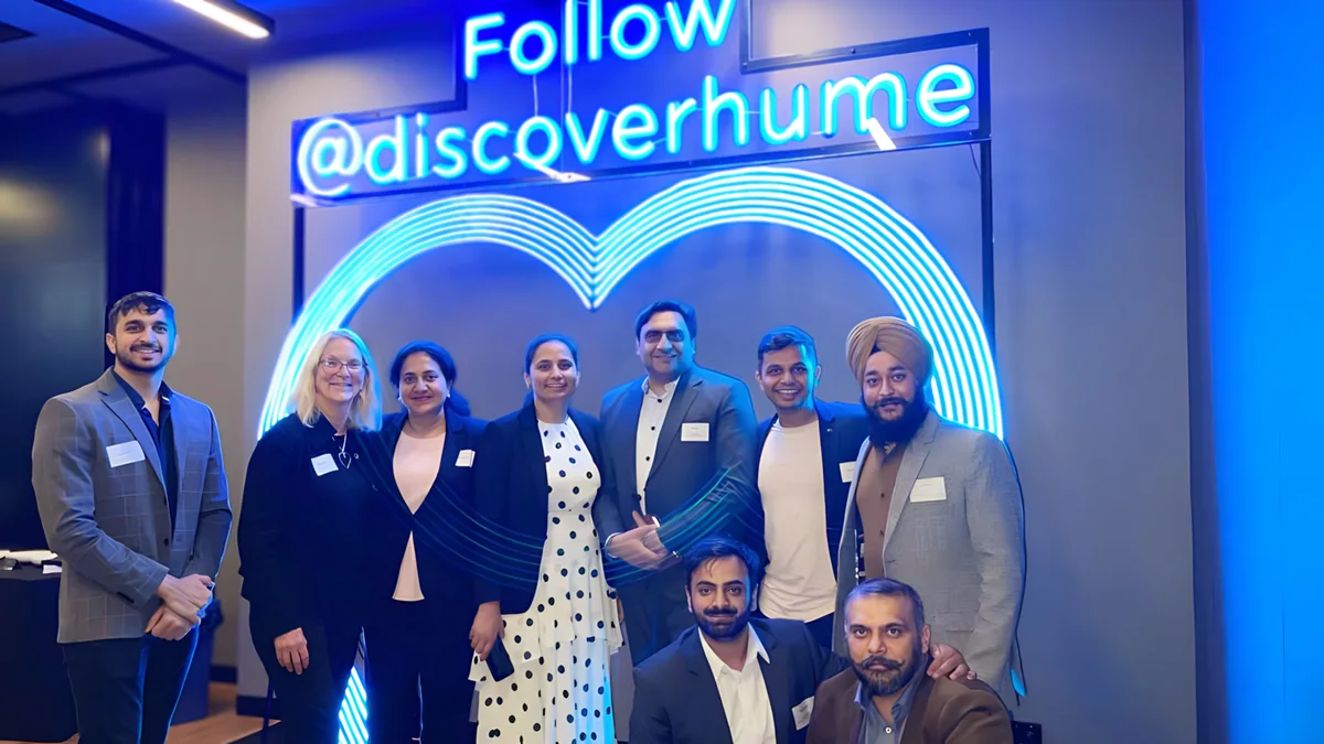 A group of people pose with a neon sign promoting the @discoverhume social media account at a business event.