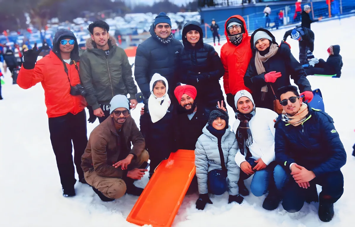 A group enjoys a snowy day outdoors, showcasing team spirit and Sukh Sandhu's involvement.